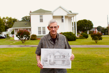 meredith pritchard man holding a drawing of the house he's posed in front of